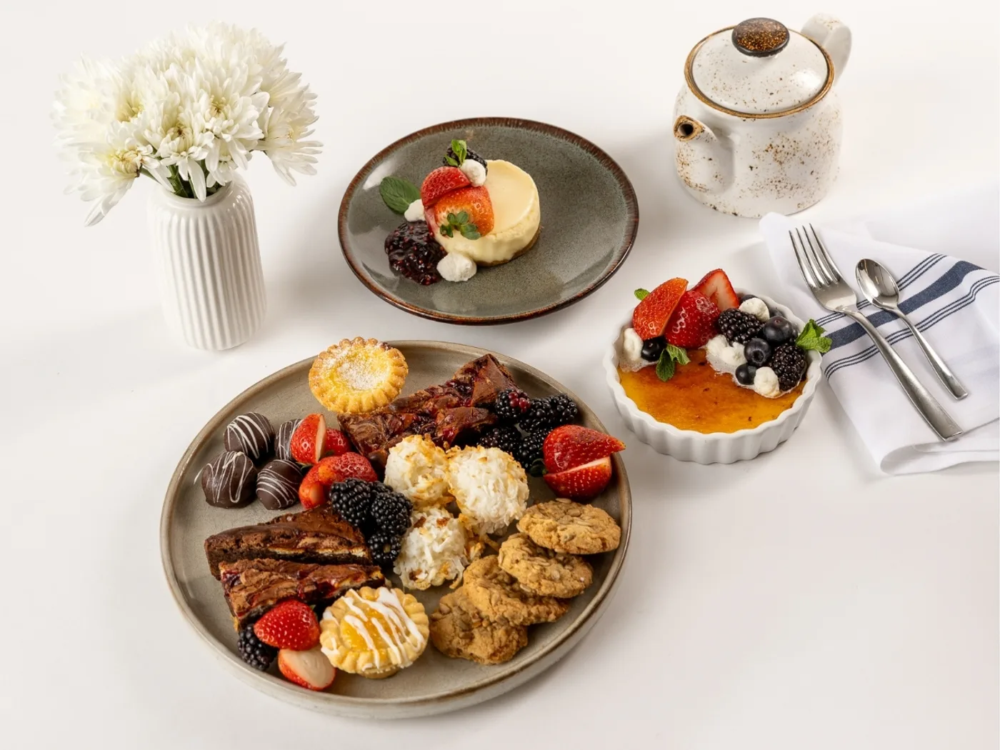 Dessert spread of chocolate truffles, tortes, macaroons, and cookies on a platter beside a berry-topped crème brûlée, cheesecake, and tea service