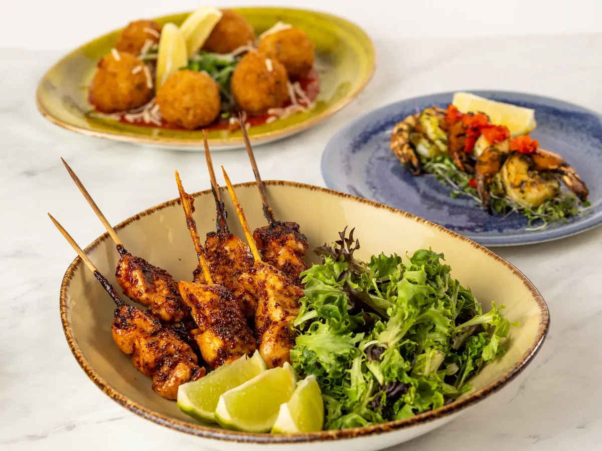 Three plates of hors d’oeuvres: crispy arancini with lemon, grilled shrimp with roasted peppers, and skewered chicken satay with mixed greens and lime