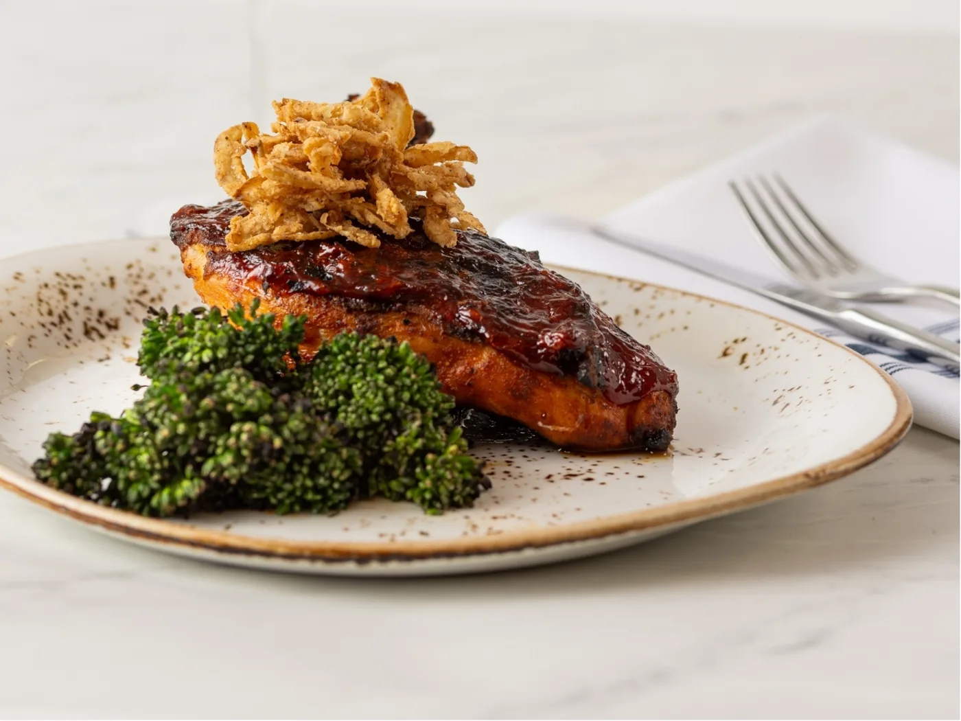 Plated glazed entrée topped with crispy onion straws and served with charred broccolini on a speckled dinner plate