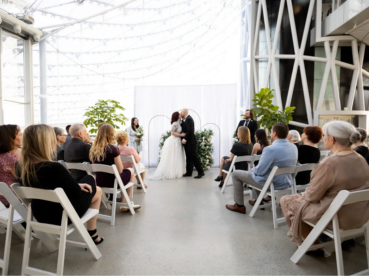 Couple sharing their first kiss at the altar during an indoor ceremony under the glass roof at The Leaf, with guests seated on white chairs on either side