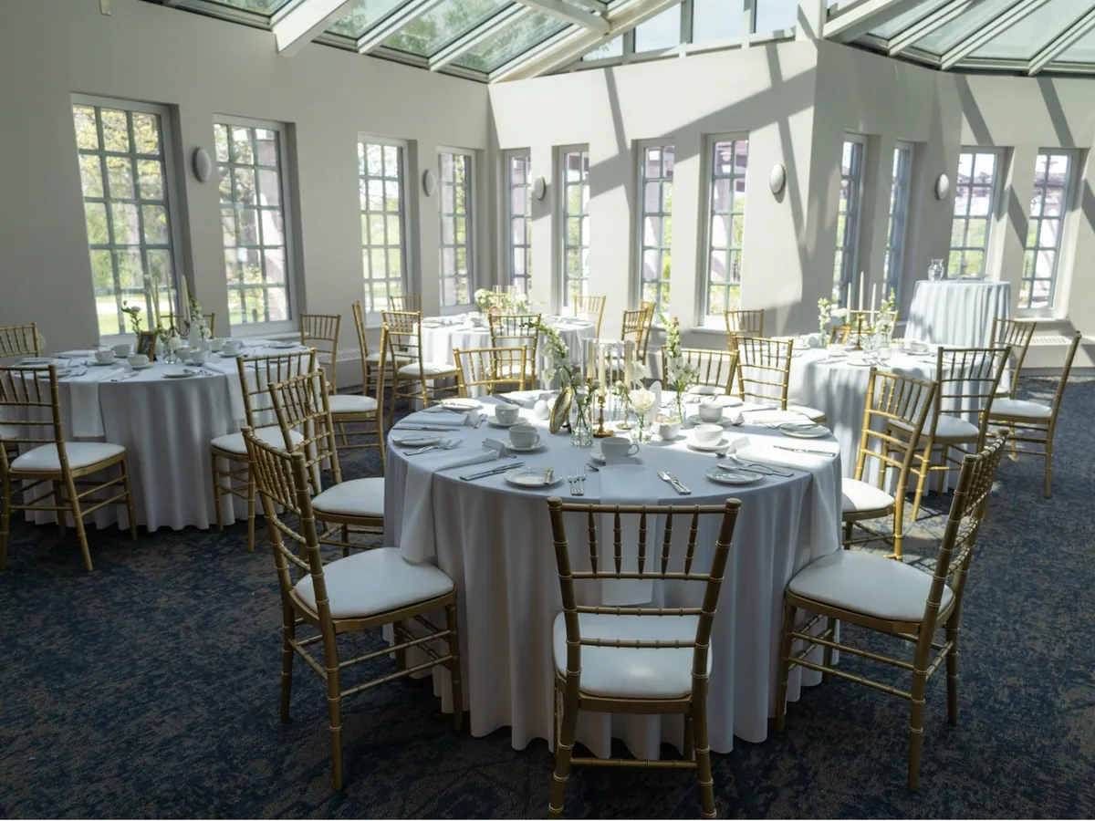 Round tables dressed in white linens with gold chiavari chairs and floral centrepieces set for a reception under the glass roof of the Pavilion Atrium